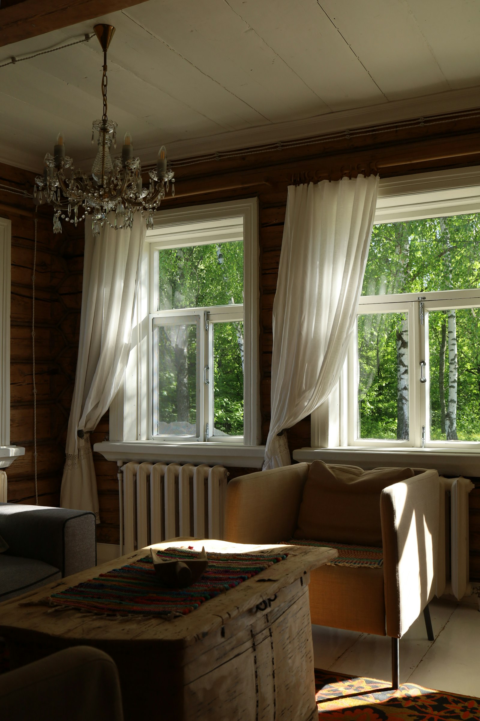 A living room with white sheer curtains at two tall windows, warm summer light and a tan armchair