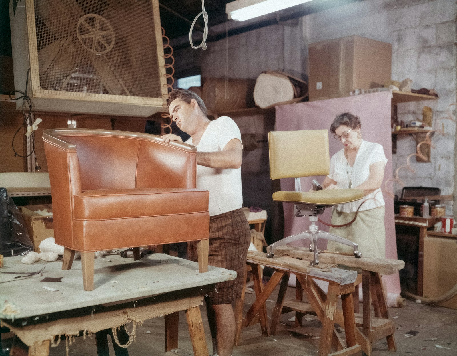 Upholsterers at work in an atelier — a tan leather tub chair on trestles being re-covered
